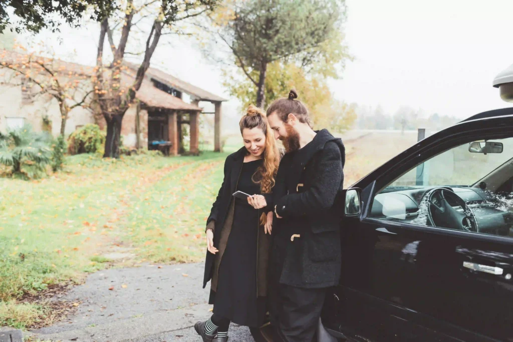 Smiling couple in black coats leans against a luxury black car, checking a smartphone. Enjoy top-rated Black Car Service Annapolis for stylish travel.