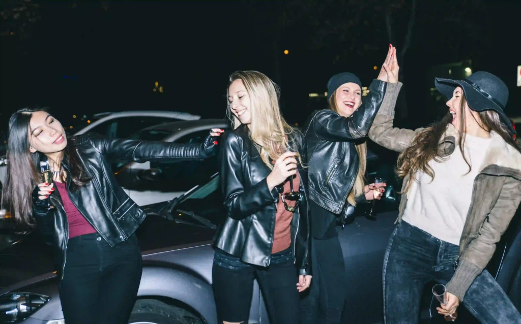 Four stylish women celebrating at night with drinks, laughing near luxury cars. Travel safely with Black Car Service Annapolis.