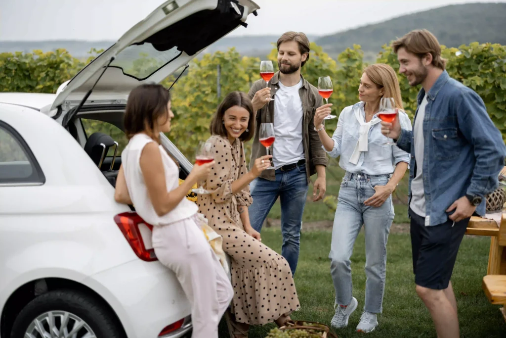 Friends enjoy wine tasting by a vineyard, toasting near a white car. Black Car Service Annapolis offers luxury transport for scenic tours.